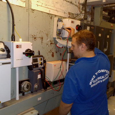 A Mike Thompson Electrical electrician in a branded blue polo shirt testing equipment in a plant room